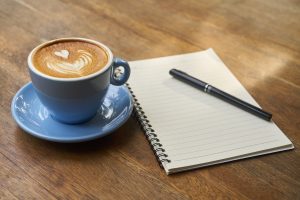 a blue cup and saucer with a coffee in on a wooden table with a blank notebook and pen next to it