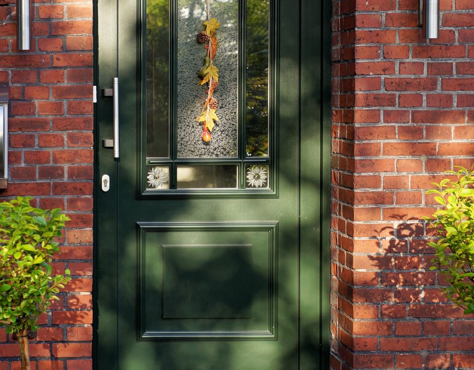 a green front door with a frosted glass panel in the middle on a red brick building with a plant either side and a doormat