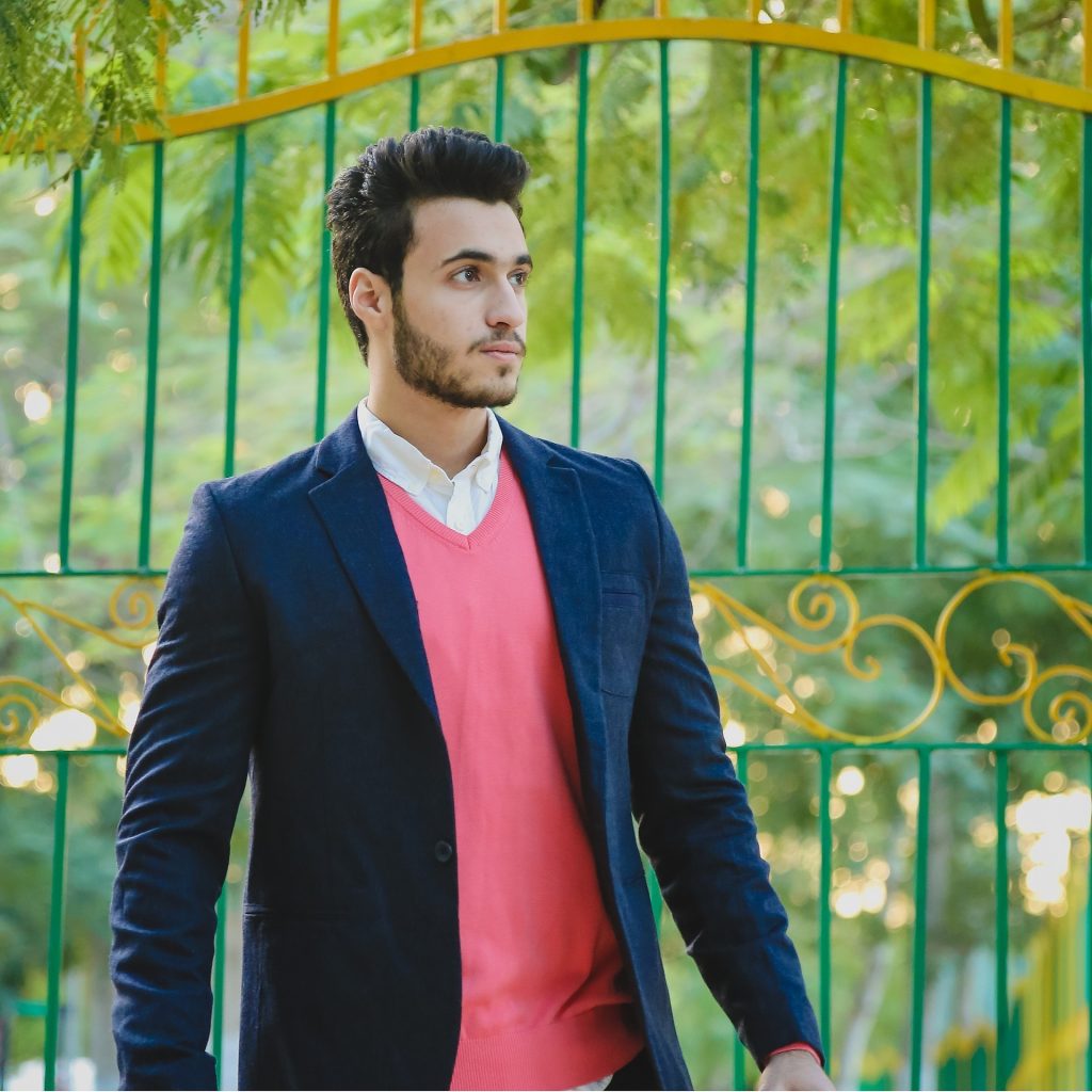 a dark haired man outside a green gate behind it is green trees. he is wearing a dark jacket and pink top