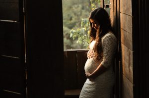 a pregnant woman holding her bump in a white dress in a dark room by an open window that has green foliage outside it