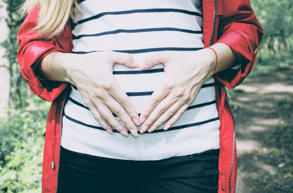 the torso of a pregnant woman. she is wearing a white and blue striped top, blue jeans and a red jacket. she is holding her hands in a heart shape over her bump