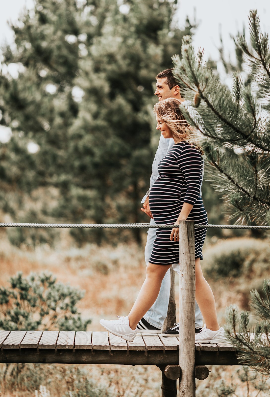 a couple walking along a wooden bridge holding hands. the woman is pregnant