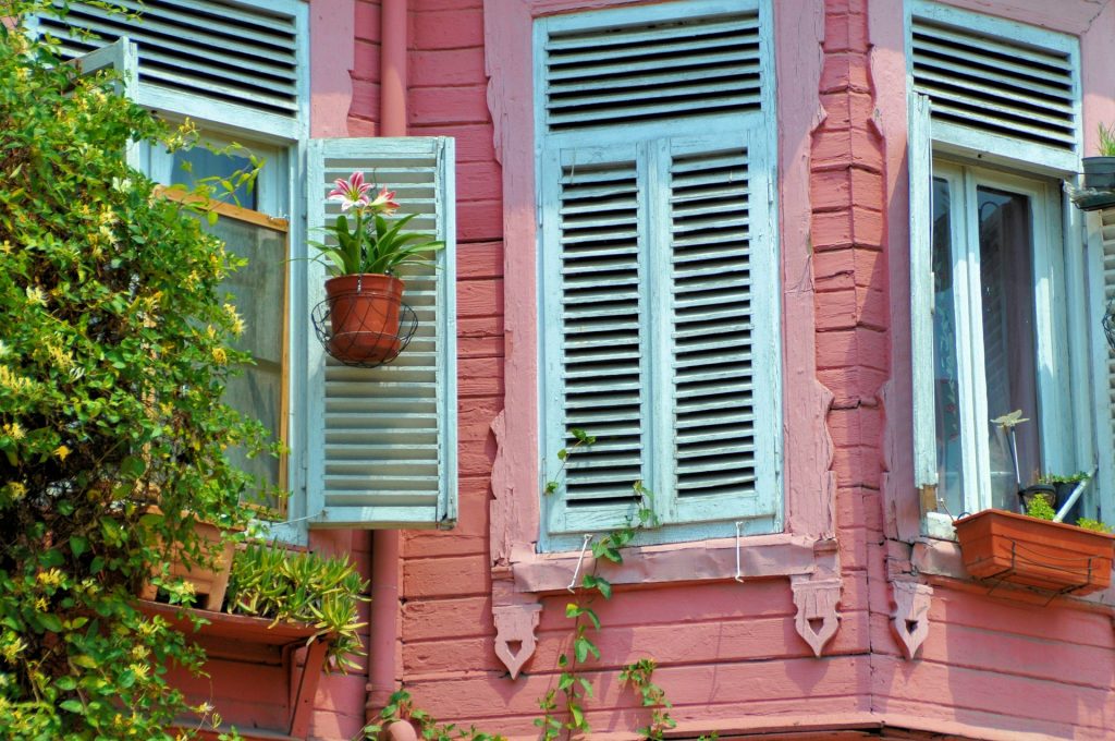 a pink house with white shuttered winders and plants around the windows