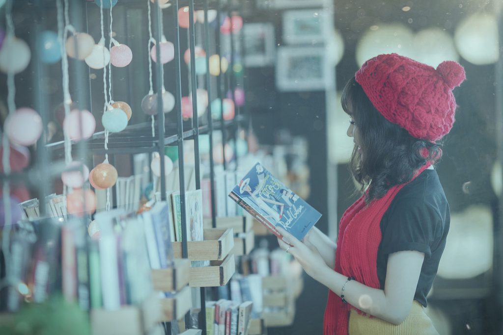 a woman stood at a wall of books reading one and laughing wearing a red hat and scarf