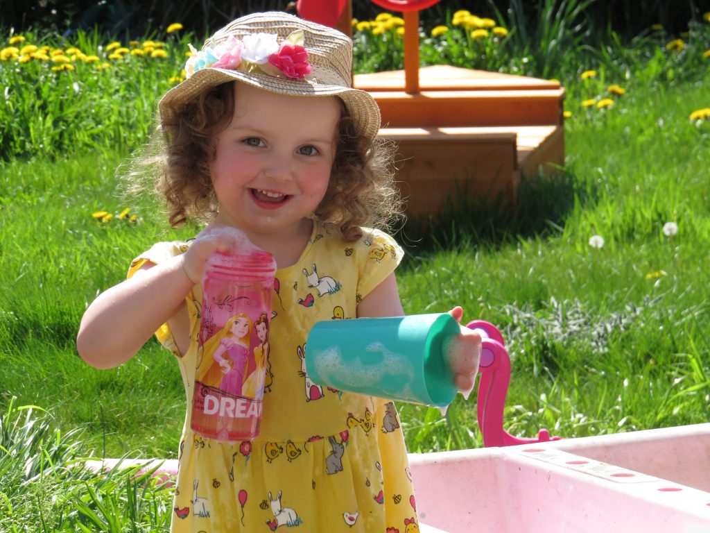 alyssa playing at a water table in a yellow dress and summer hat smiling at the camera