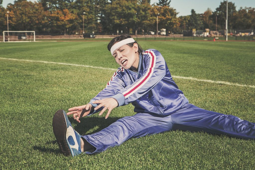 a woman in a blue tracksuit sat on grass with her legs out trying to touch her toes