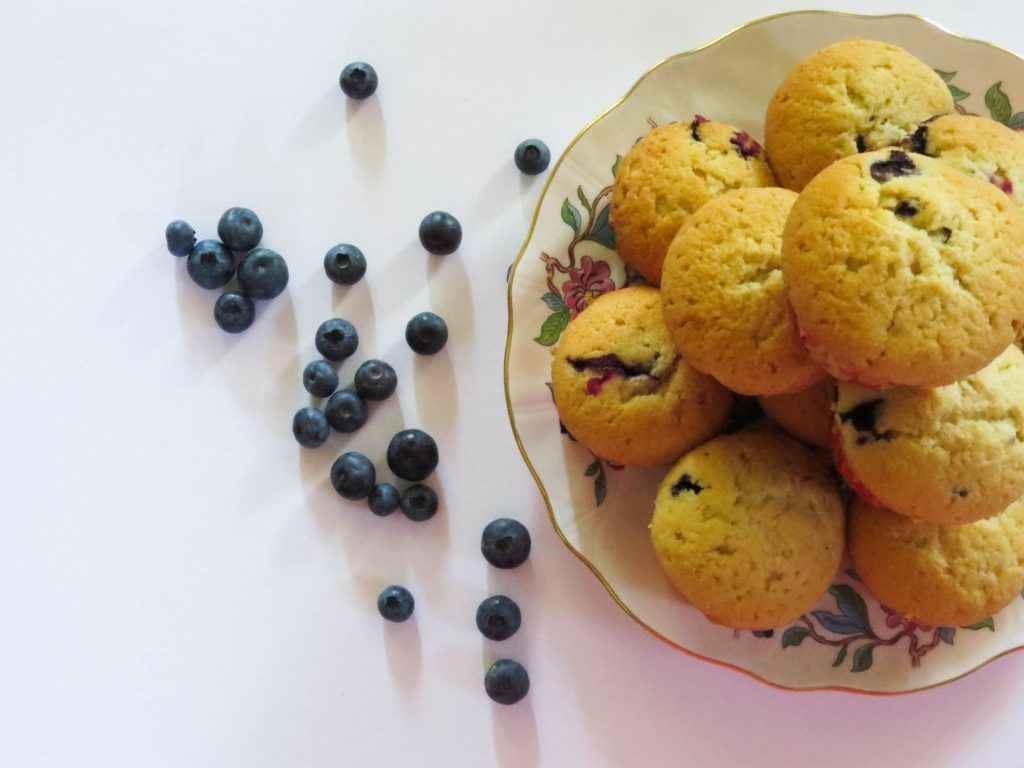 a white background and on it a plate of blueberry muffins stacked up with loose fresh blueberries next to them