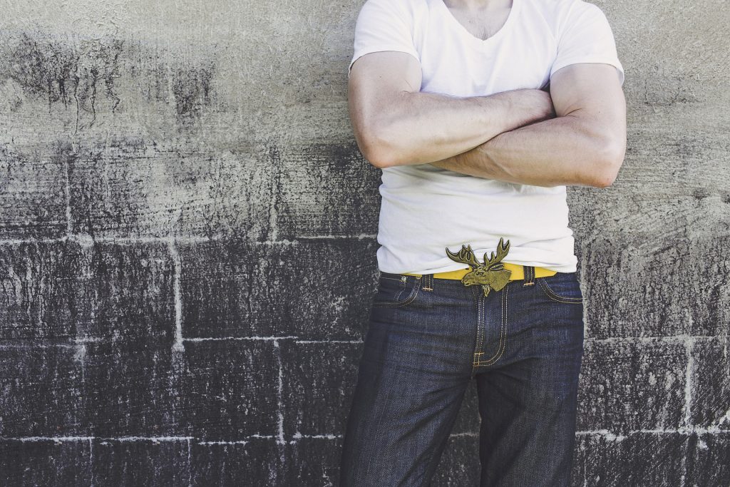 a man leaning against a wall with folded arms wearing jeans and a white tshirt