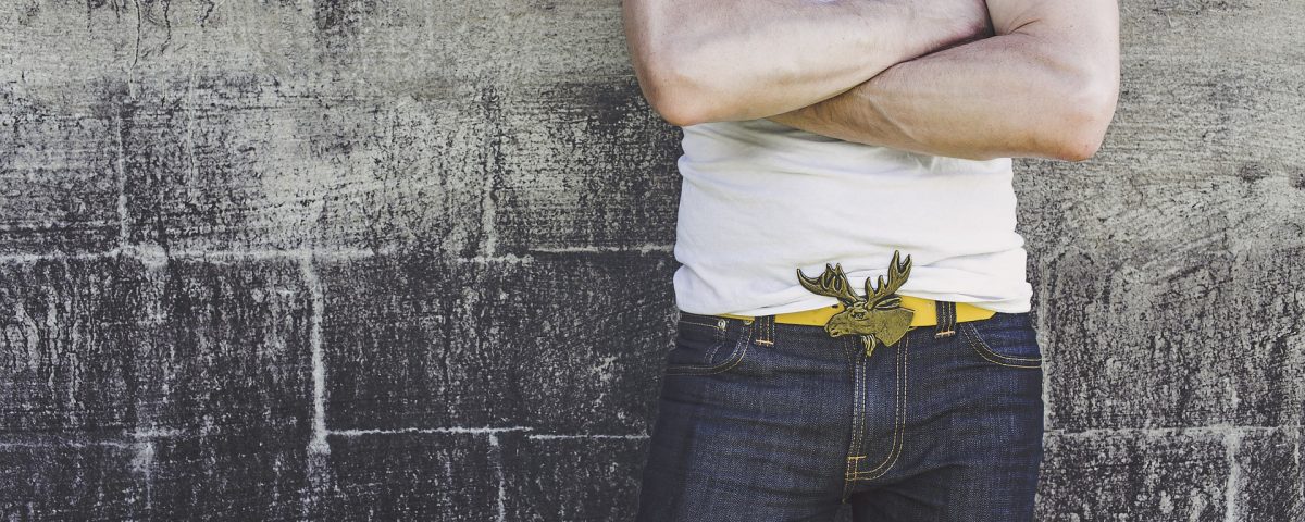 a man leaning against a wall with folded arms wearing jeans and a white tshirt