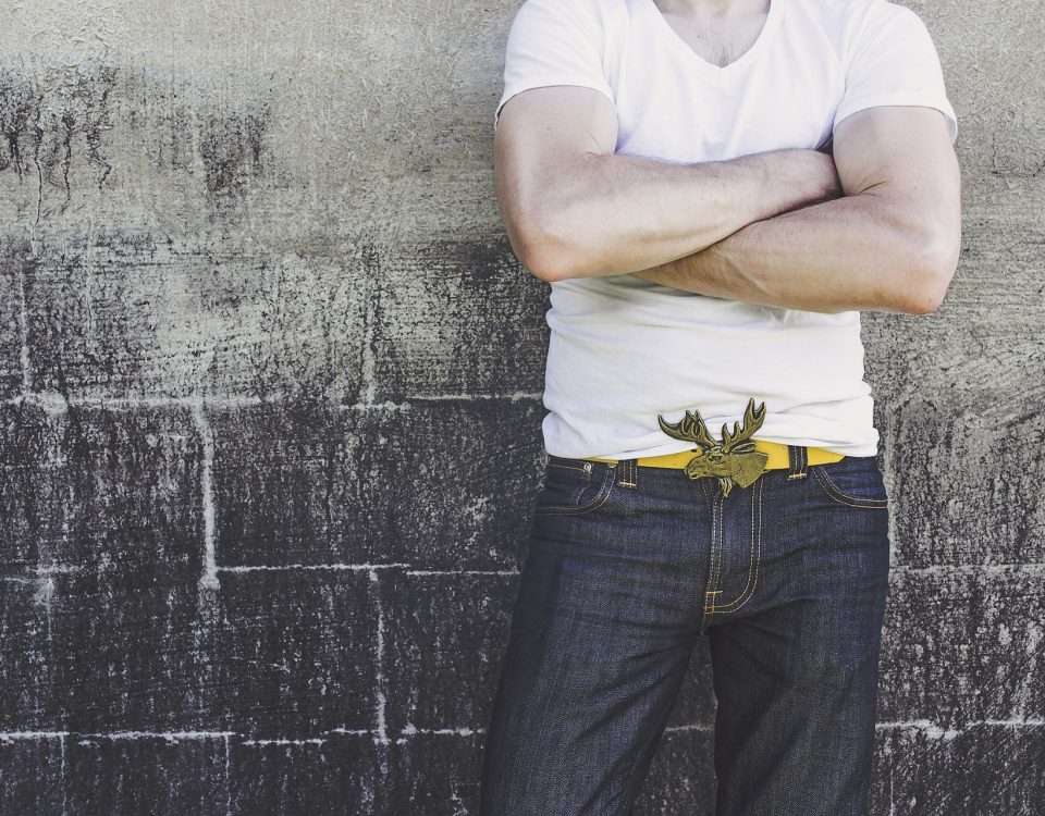 a man leaning against a wall with folded arms wearing jeans and a white tshirt